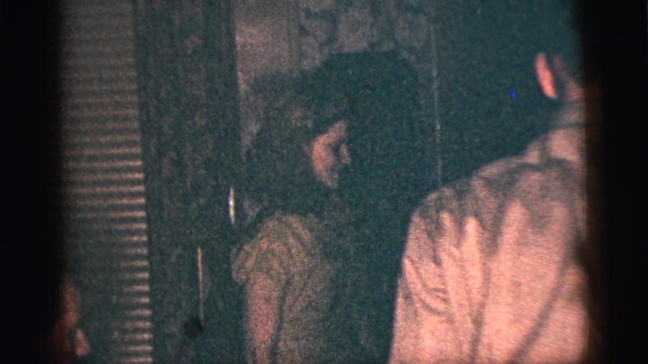 In supported 1956 New York City home-movie footage, a young woman in a light dress stands near relatives in a dim interior.