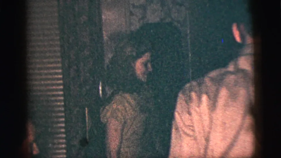 In supported 1956 New York City home-movie footage, a young woman in a light dress stands near relatives in a dim interior.