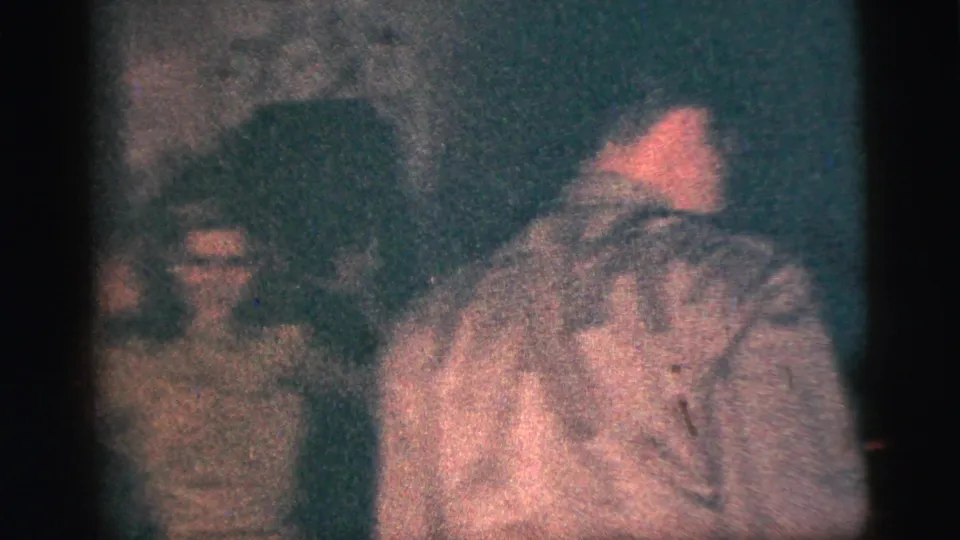 Several people appear in a dim 1956 New York City home-movie interior, including a man in a light shirt facing partly away.