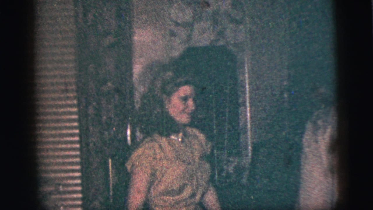 A young woman in a light dress smiles and raises a hand toward the camera in supported 1956 New York City home-movie footage.