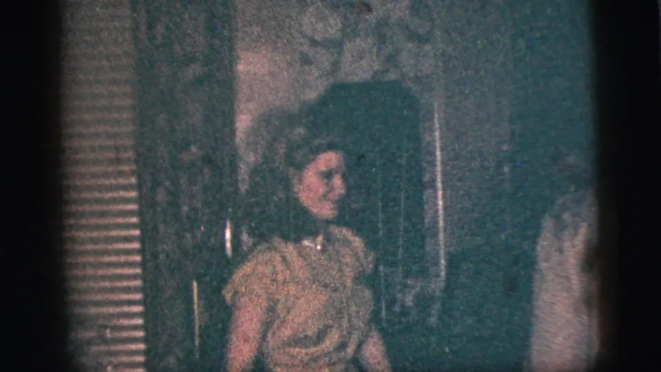 A young woman in a light dress smiles and raises a hand toward the camera in supported 1956 New York City home-movie footage.
