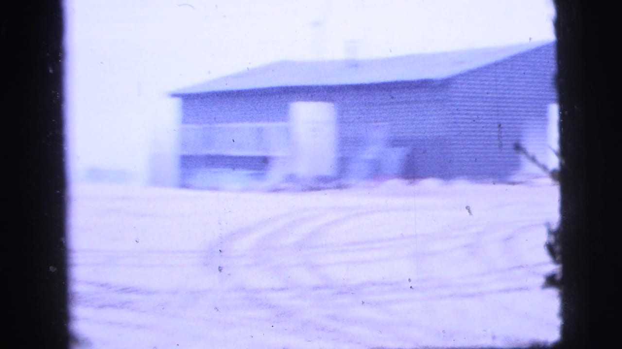 Blue-tinted 8mm frame of a low industrial building across an open yard or road in New York City, dated by clip metadata to 1956.