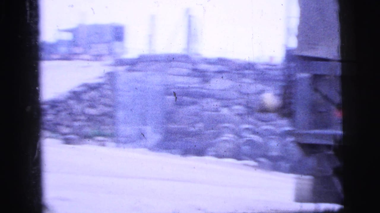 1956 New York City home-movie frame showing stacked stone or masonry and a small structure beside an industrial roadside.