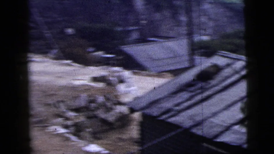 Handheld 1956 New York City 8mm frame showing rubble, exposed earth, corrugated siding, and dark vertical foreground shapes.