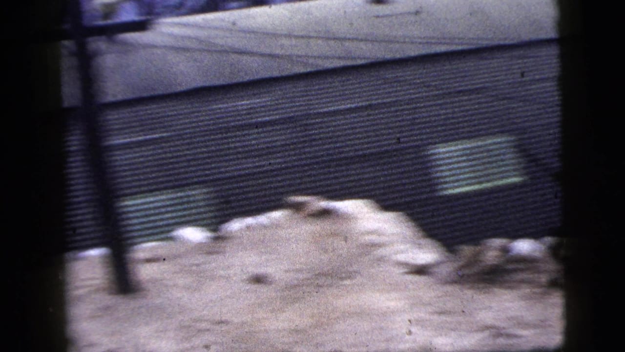1956 New York City 8mm frame showing a corrugated metal wall, debris, bare ground, and blurred dark foreground obstructions.