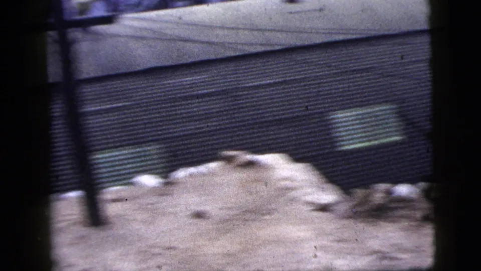 1956 New York City 8mm frame showing a corrugated metal wall, debris, bare ground, and blurred dark foreground obstructions.