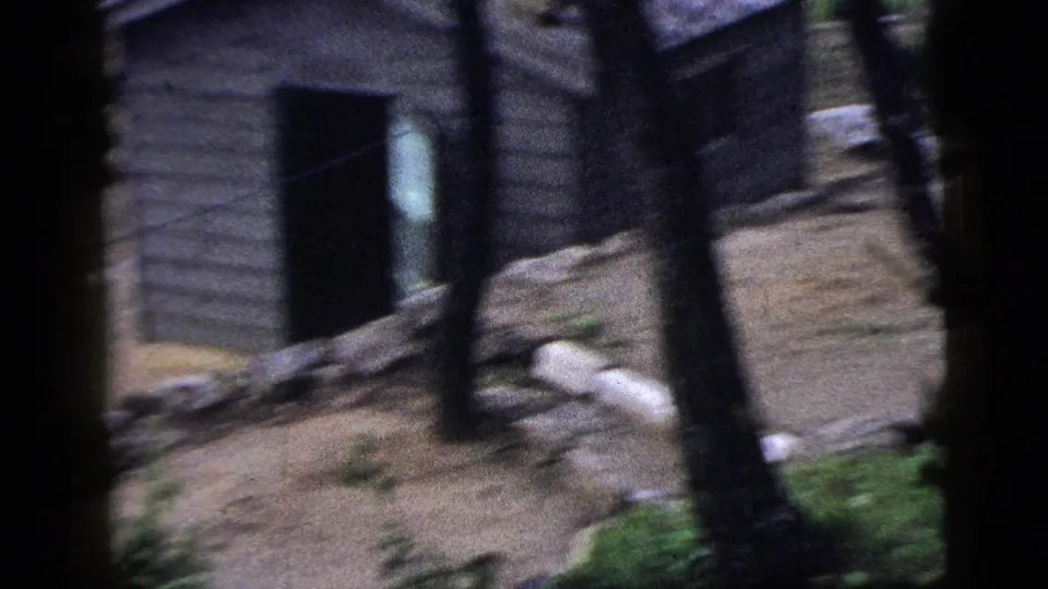 Handheld 1956 New York City frame showing a weathered wall, dark doorway, debris at ground level, and sparse greenery.