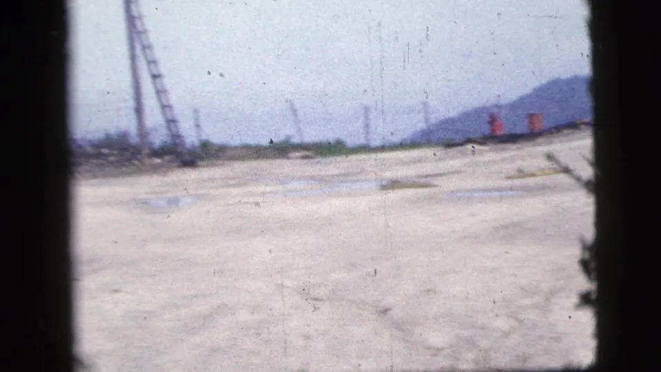 1956 New York City 8mm frame looking across cracked concrete, puddles, ladders, utility poles and a distant low hill.