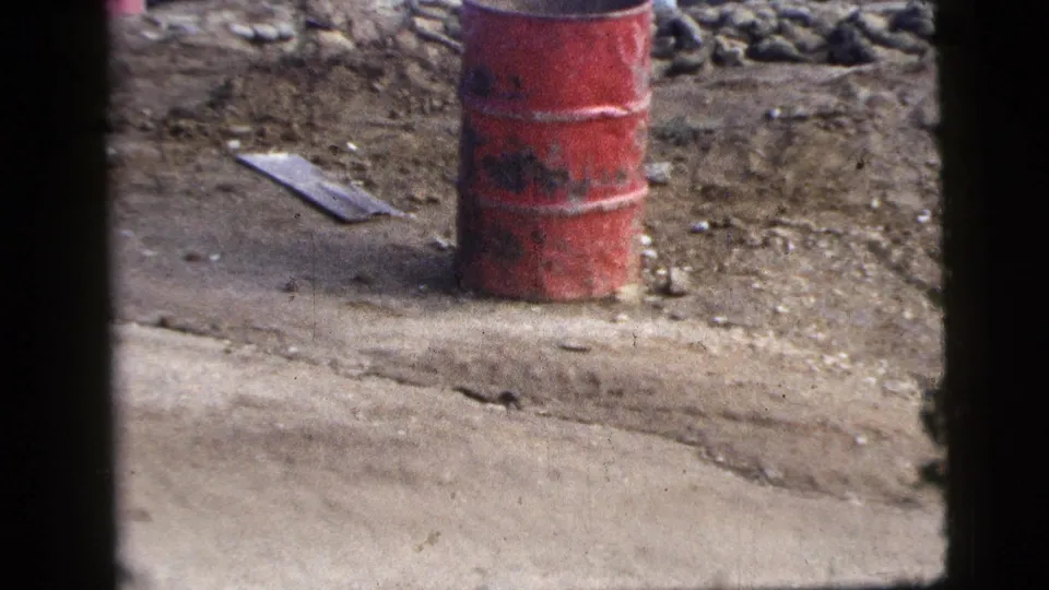 1956 New York City 8mm close view of a red steel barrel on rough ground beside rocks, debris and a concrete edge.
