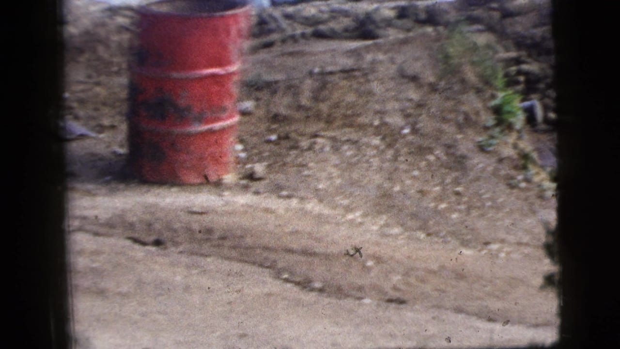 1956 New York City home-movie frame looking through a vehicle window toward a red barrel, dirt road, stone wall, and scattered debris.
