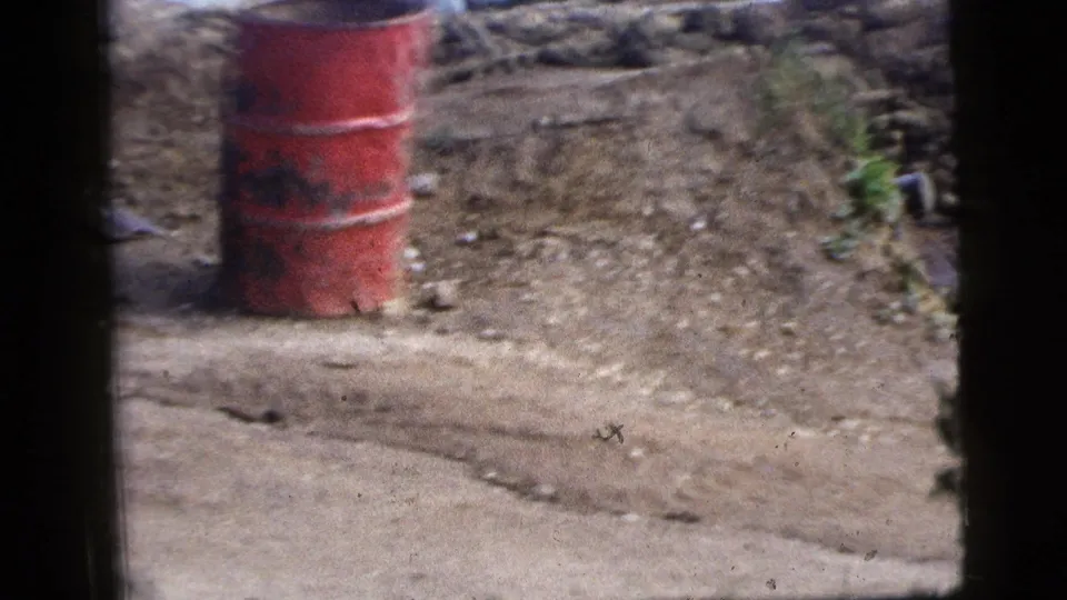 1956 New York City home-movie frame looking through a vehicle window toward a red barrel, dirt road, stone wall, and scattered debris.