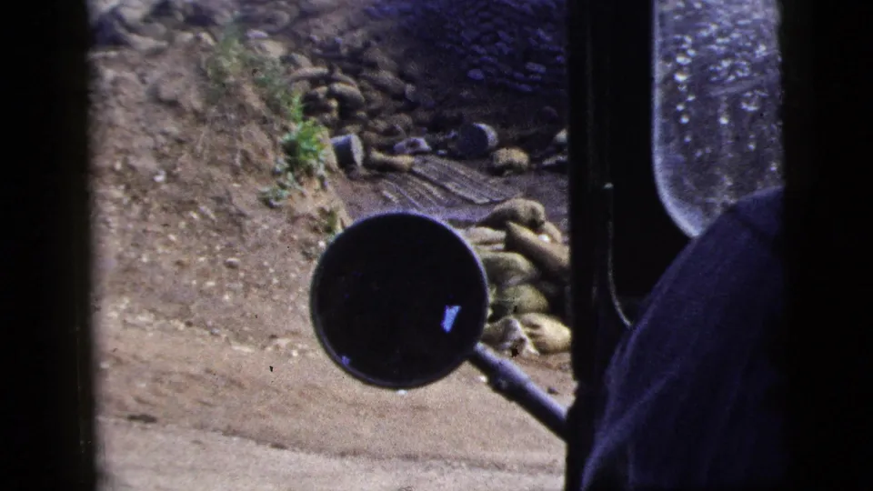 1956 New York City frame from inside a moving vehicle, showing a side mirror, window edge, dirt surface, stone wall, and debris.