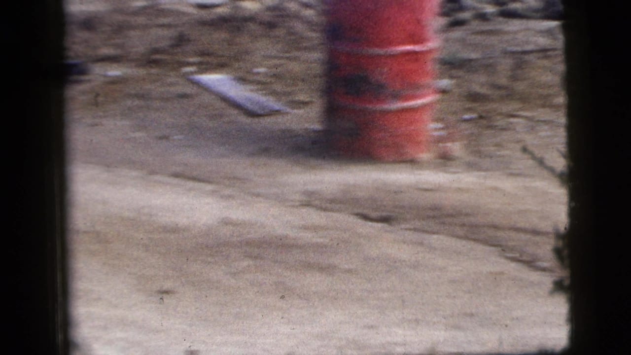 Motion-blurred 1956 New York City roadside frame with a red barrel, rough ground, debris, and vehicle-window borders.