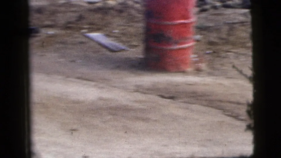 Motion-blurred 1956 New York City roadside frame with a red barrel, rough ground, debris, and vehicle-window borders.