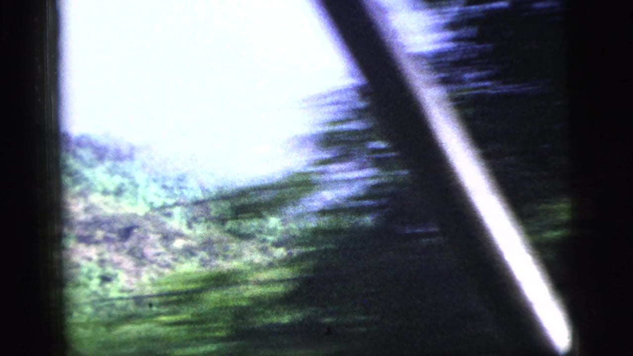 1956 8mm frame showing bright sky, blurred green foliage, and dark diagonal foreground shapes during a bumpy ride.