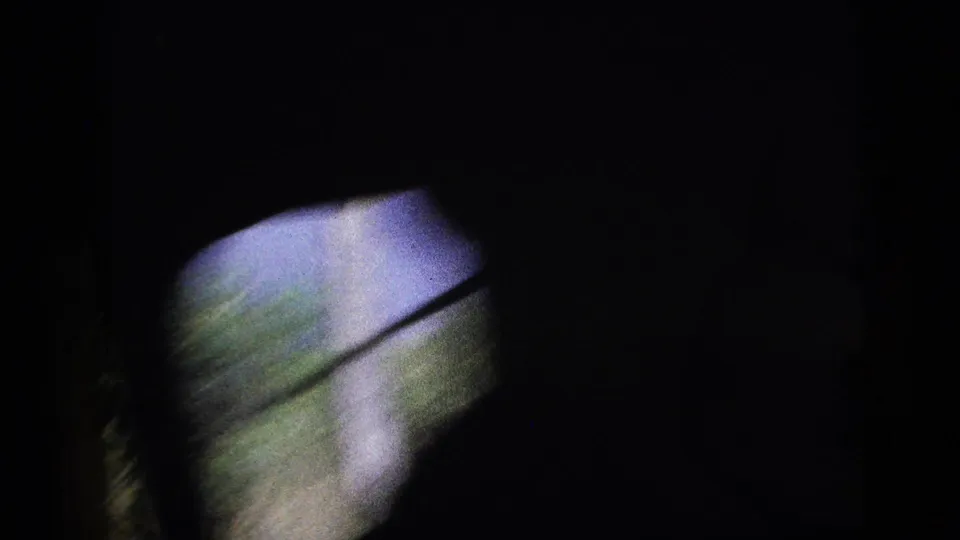 Dark in-vehicle view with a small bright window area, blurred greenery, and a diagonal frame or handle across the opening.