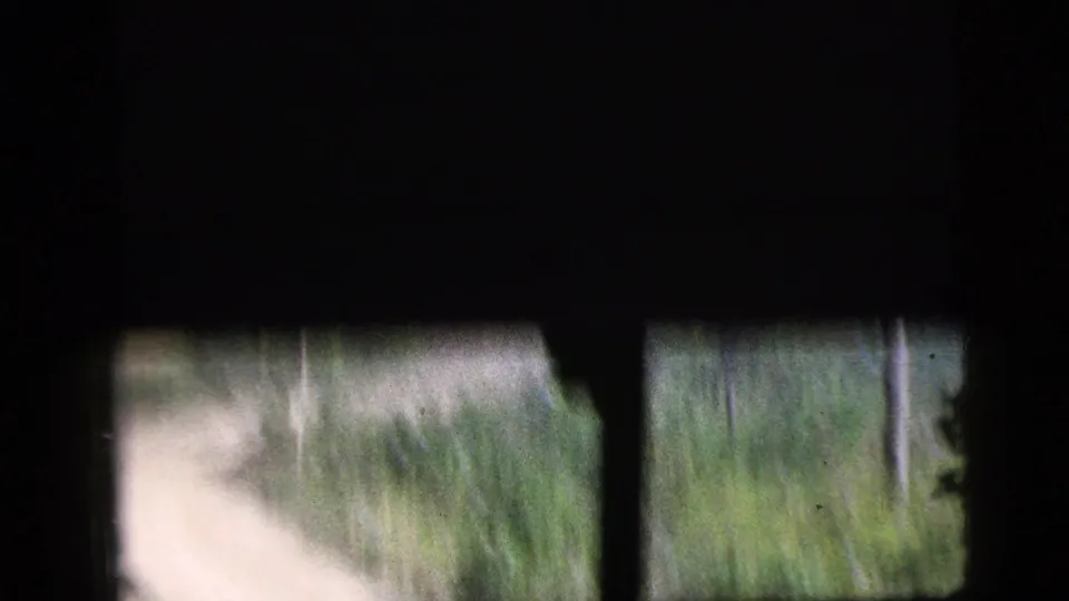 Obstructed 1956 8mm travel frame showing dark vehicle framing, a blurred pale road, and green vegetation near the lower edge.