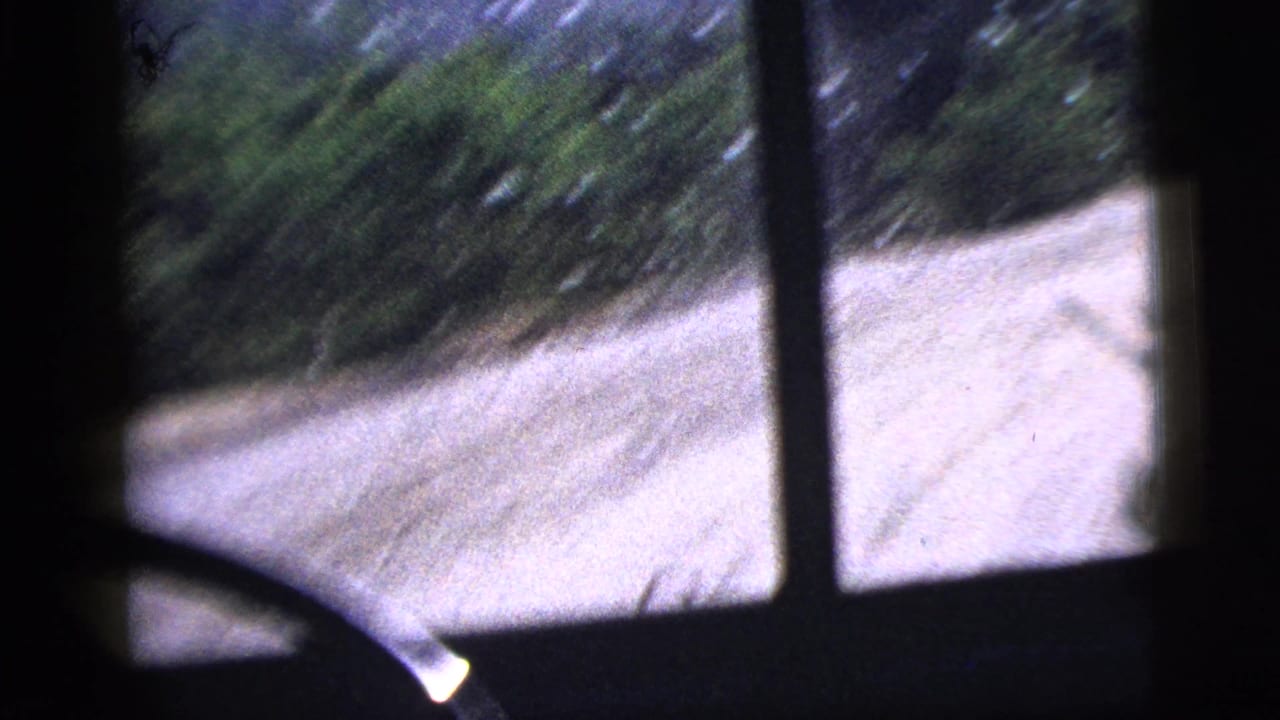 1956 8mm frame showing a pale road or path and blurred trees through dark vehicle window framing.