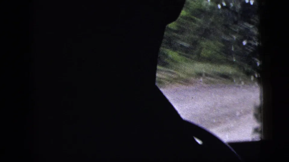 1956 8mm frame showing a driver in silhouette inside a moving vehicle, with blurred road and green roadside visible beyond the window.