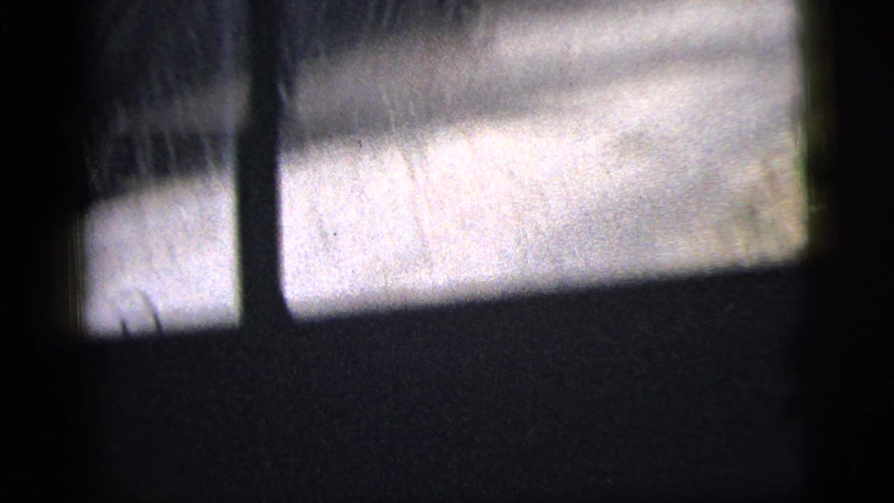 1956 8mm frame with a blurred side-window view of a pale road, dark vertical window framing, and indistinct roadside structures or posts.