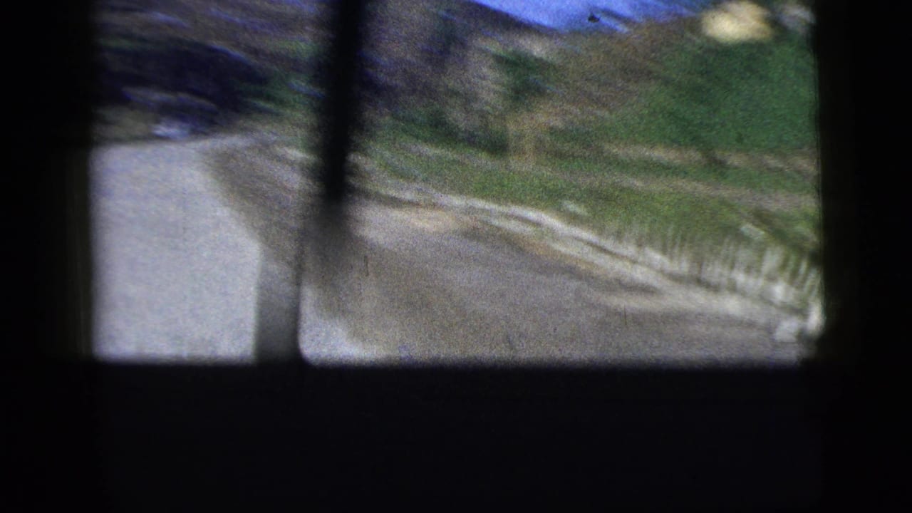 Shaky 1956 8mm view from inside a moving car, with road surface, grass verge, and dark window bars visible.