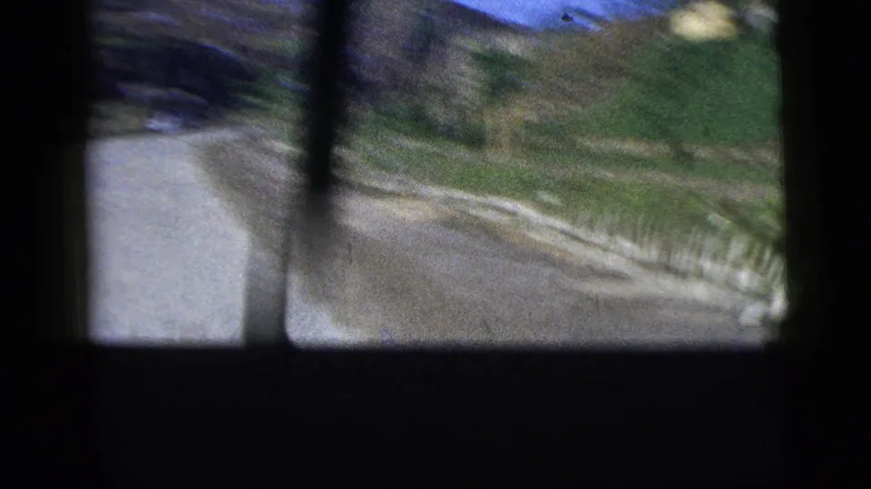 Shaky 1956 8mm view from inside a moving car, with road surface, grass verge, and dark window bars visible.