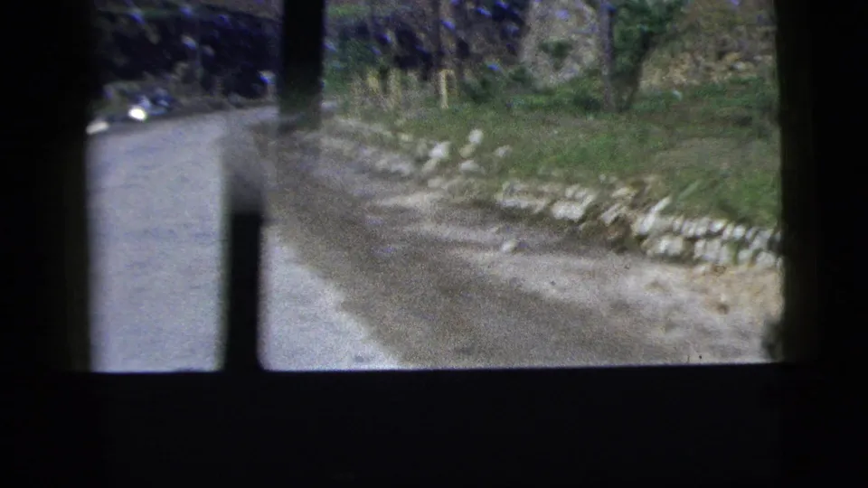 1956 8mm frame showing a narrow rural road, grassy roadside, and stone edging from a moving car viewpoint.