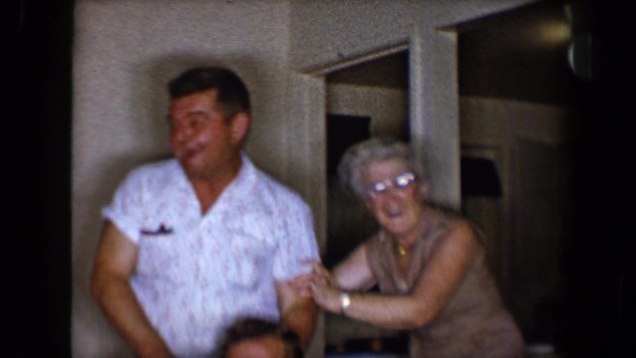 A man in a white patterned shirt stands beside an older woman with glasses in a Tucson living room, 1958 metadata supplied.