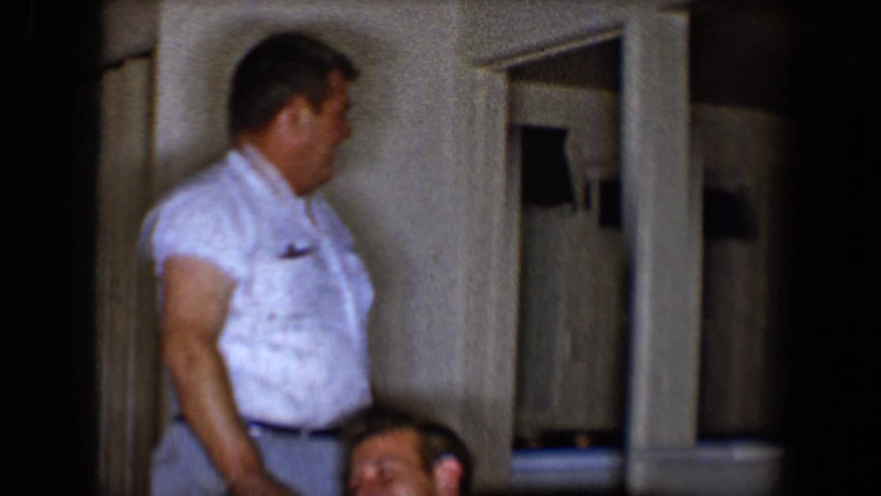 A man in a short-sleeve white shirt stands near the sofa while another man laughs at the bottom of the frame.