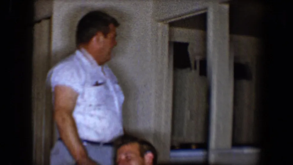 A man in a short-sleeve white shirt stands near the sofa while another man laughs at the bottom of the frame.