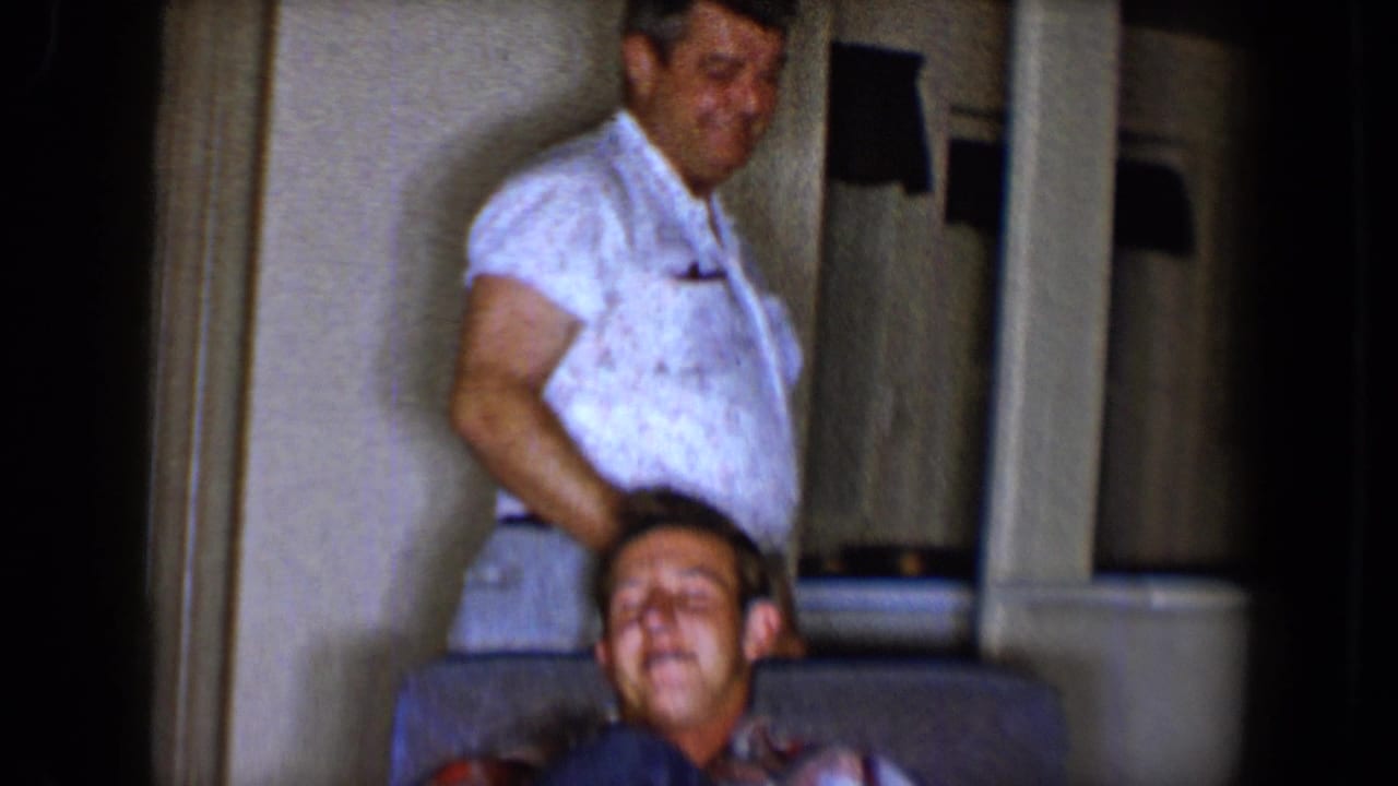 A standing man in a white shirt appears to steady a laughing seated man on a sofa in the 1958 Tucson home interior.