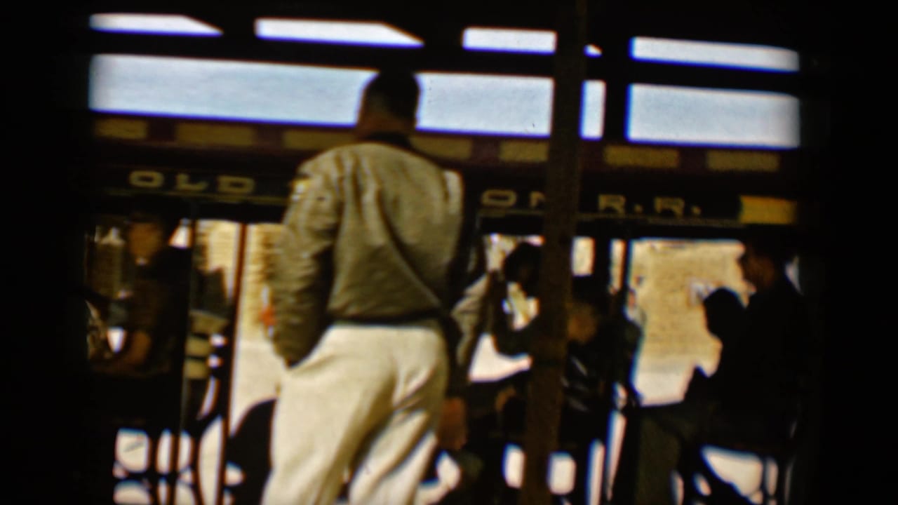 Passengers stand and sit inside an open-air trolley with partial railroad signage above them.
