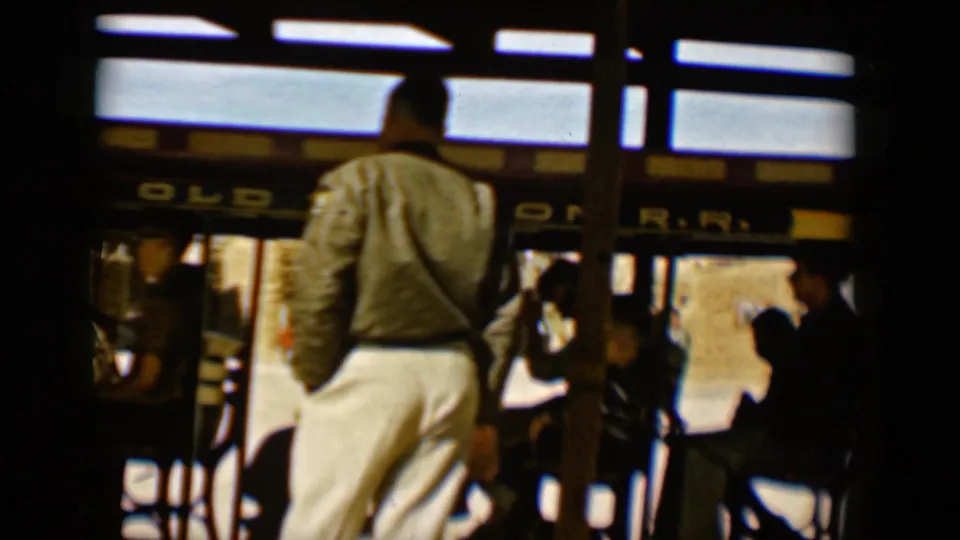 1958 Tucson home-movie frame showing adults boarding or riding an open-air Old Tucson R.R. trolley, with partial signage and wooden posts visible.