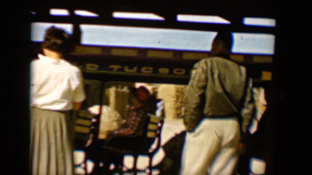 A woman in a white blouse stands near seated riders inside the open trolley car.