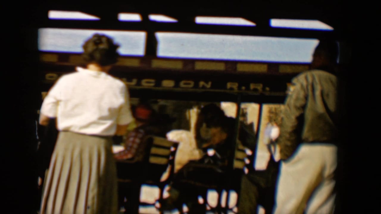 Home-movie frame from the 1958 Tucson trolley scene, with a woman in a skirt, a man in white trousers, seated riders, and partial Old Tucson R.R. lettering.