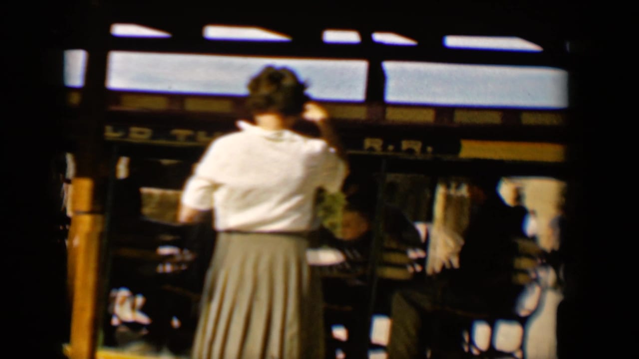 Final 1958 Tucson frame showing the open-air trolley from a shaded angle, with a standing woman, riders, vertical posts, and partial railroad lettering.