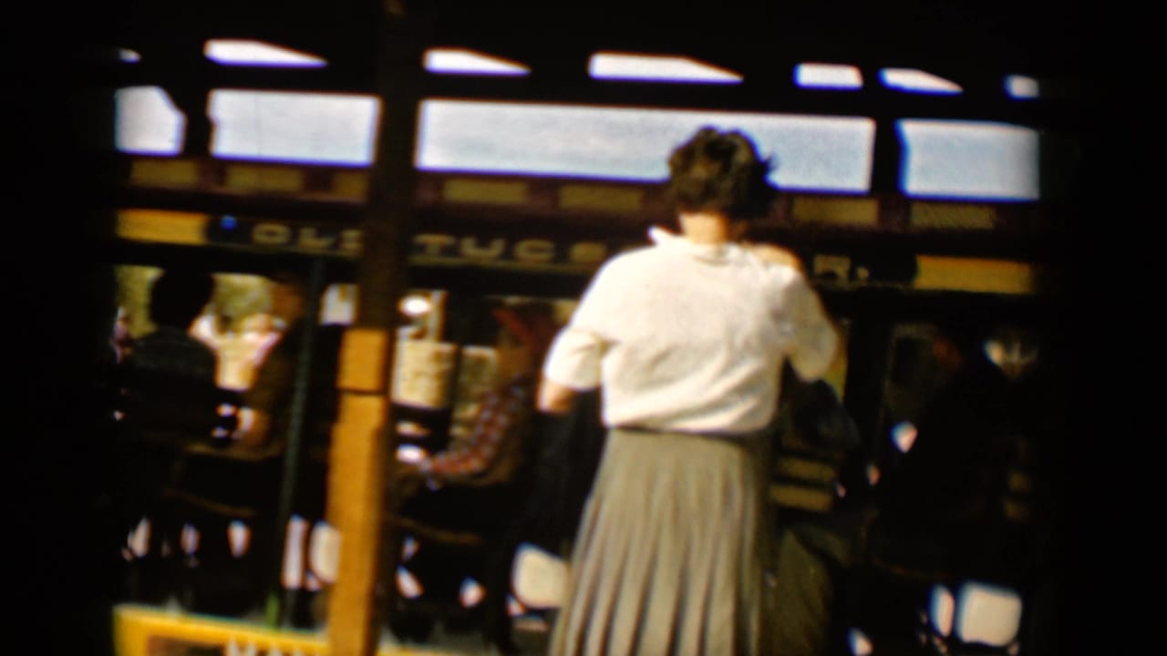 Woman in a white blouse stands near an open-sided trolley with seated passengers inside.