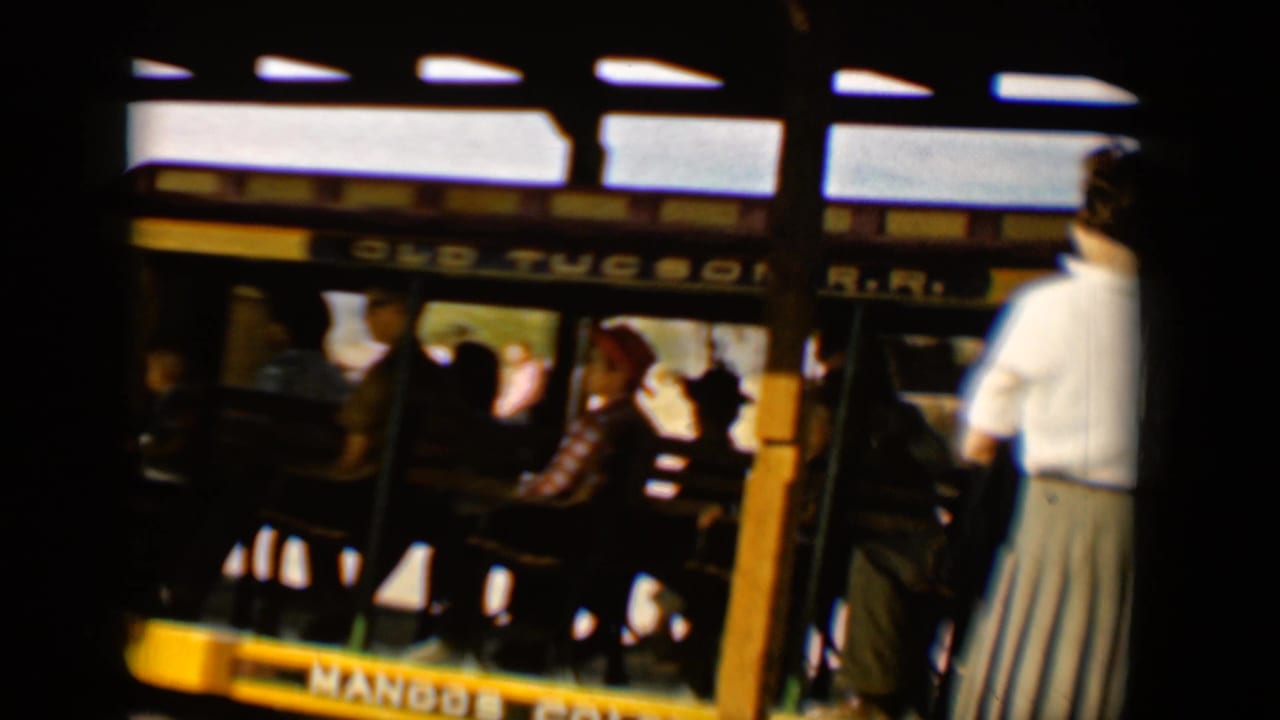 Old Tucson R.R. trolley car in Tucson, 1958, with seated riders visible under the roof and a standing figure at the edge.