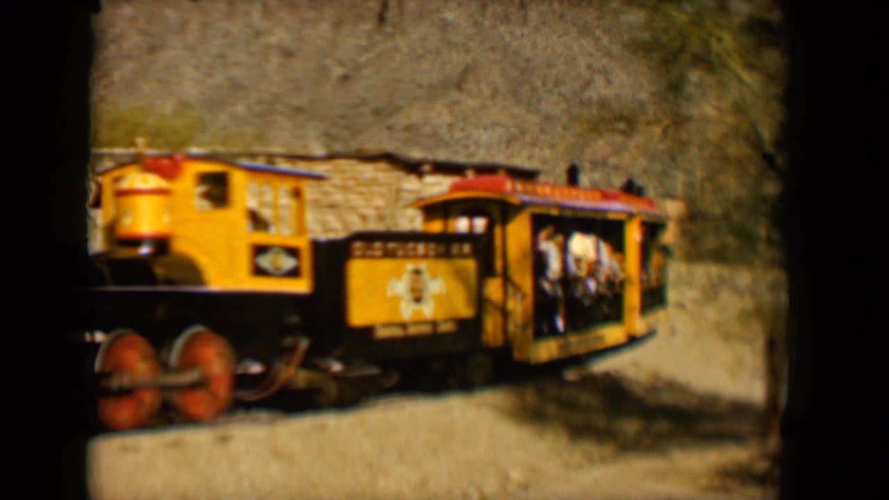 The train passes close to the camera, showing a yellow open-sided car with riders inside in Tucson, 1958.