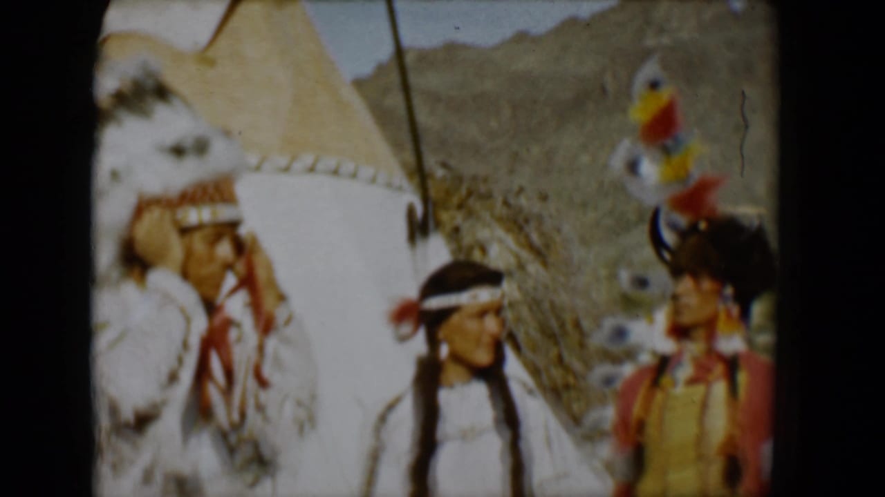 Three adults in Native American-style attire stand near a tipi in a desert setting.