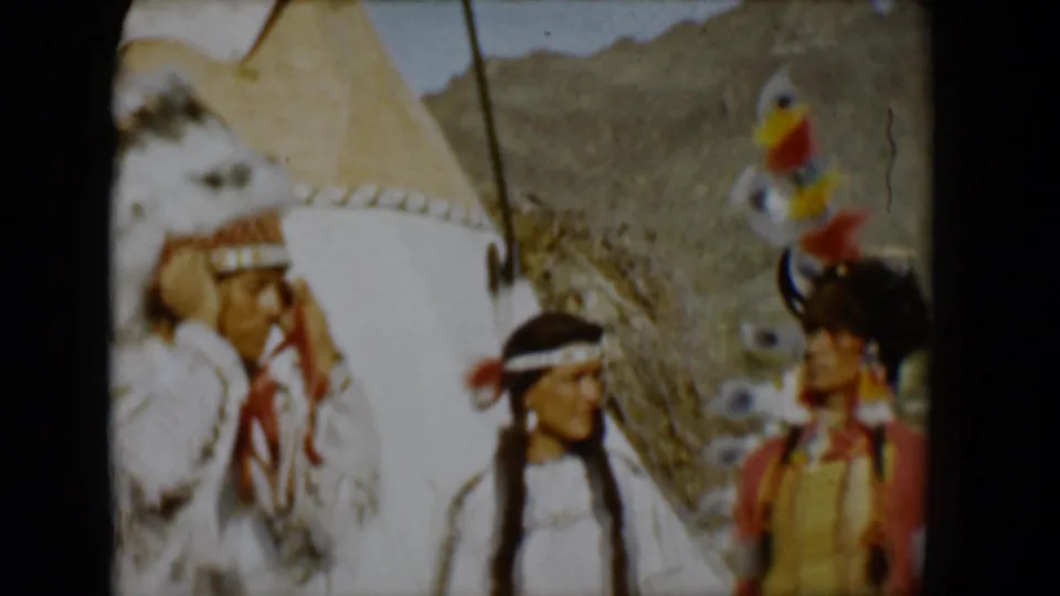 1958 Tucson home-movie frame showing three adults in Native American-style dress near a tipi, with feathered headdresses, fringed clothing, and desert hills behind them.