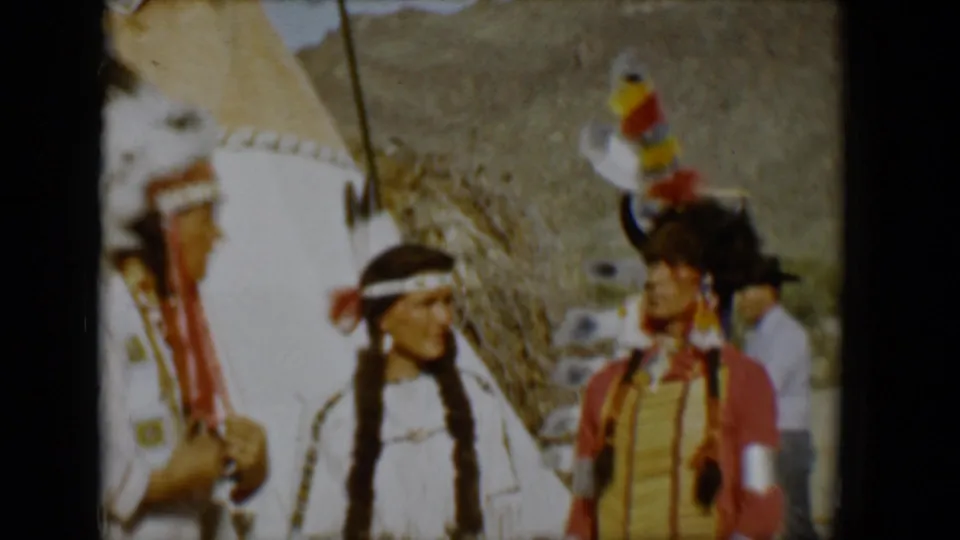 1958 Tucson 8mm frame with two adults in Native American-style clothing facing each other beside a tipi, with feathers, braids, fringe, and sunlit desert background visible.