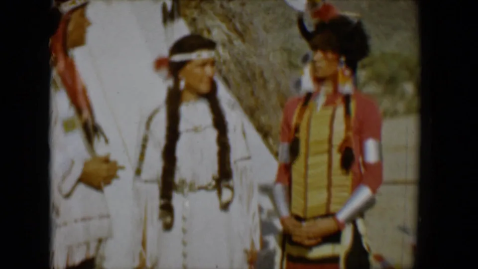 1958 Tucson home-movie frame showing two adults in Native American-style dress near a tipi, including a white fringed garment, long braids, and a red-and-yellow outfit.