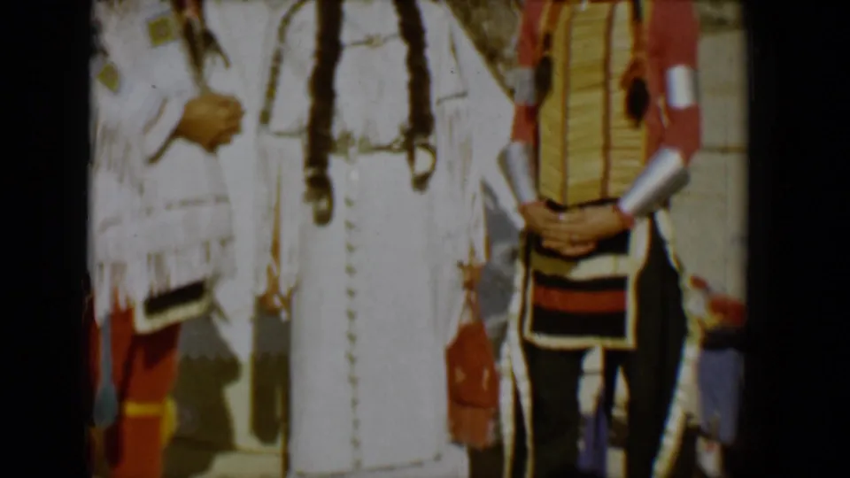 1958 Tucson 8mm frame showing two adults posed near a tipi in Native American-style attire, with a feathered headdress, braids, fringe, and desert hills in the background.