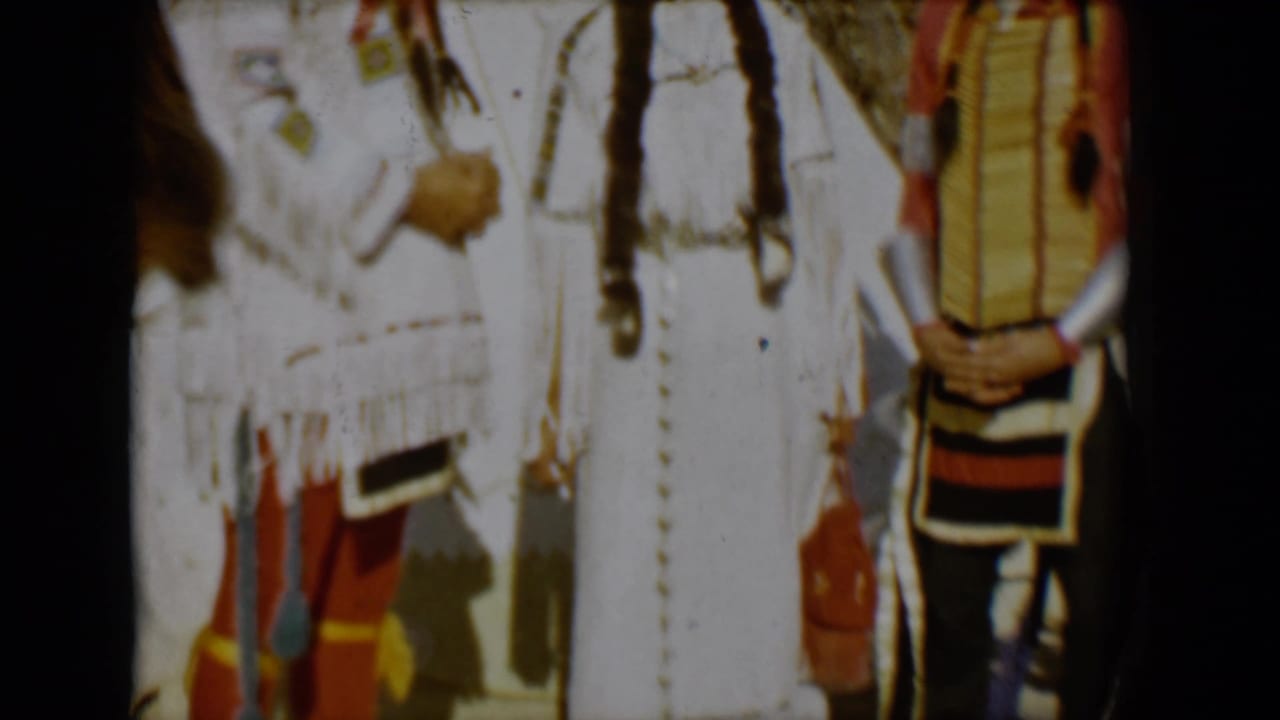 1958 Tucson home-movie frame showing people in feathered regalia and fringed garments near a teepee, with heavy exposure and frame edge shadows.
