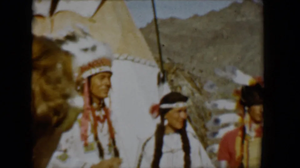 1958 Tucson home-movie frame showing several figures in regalia near a teepee, with desert mountains visible beyond the scene.