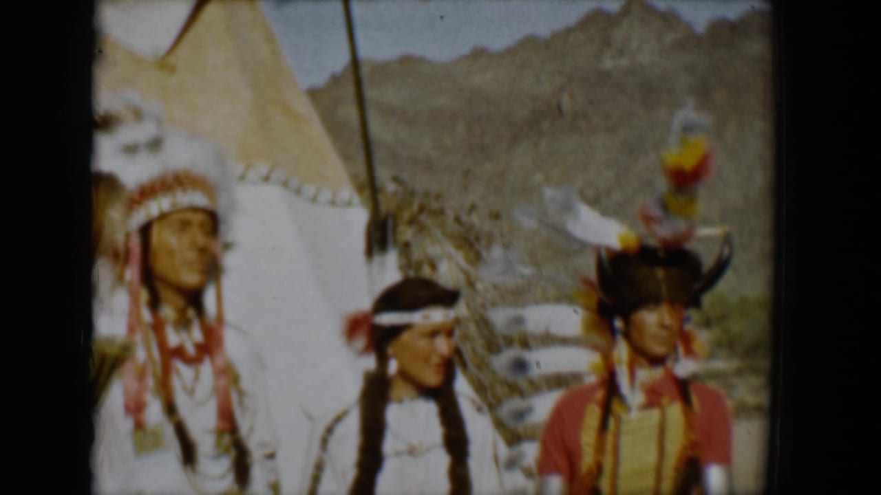 1958 Tucson-area 8mm frame showing three people in regalia posed beside a canvas teepee against a desert mountain backdrop.