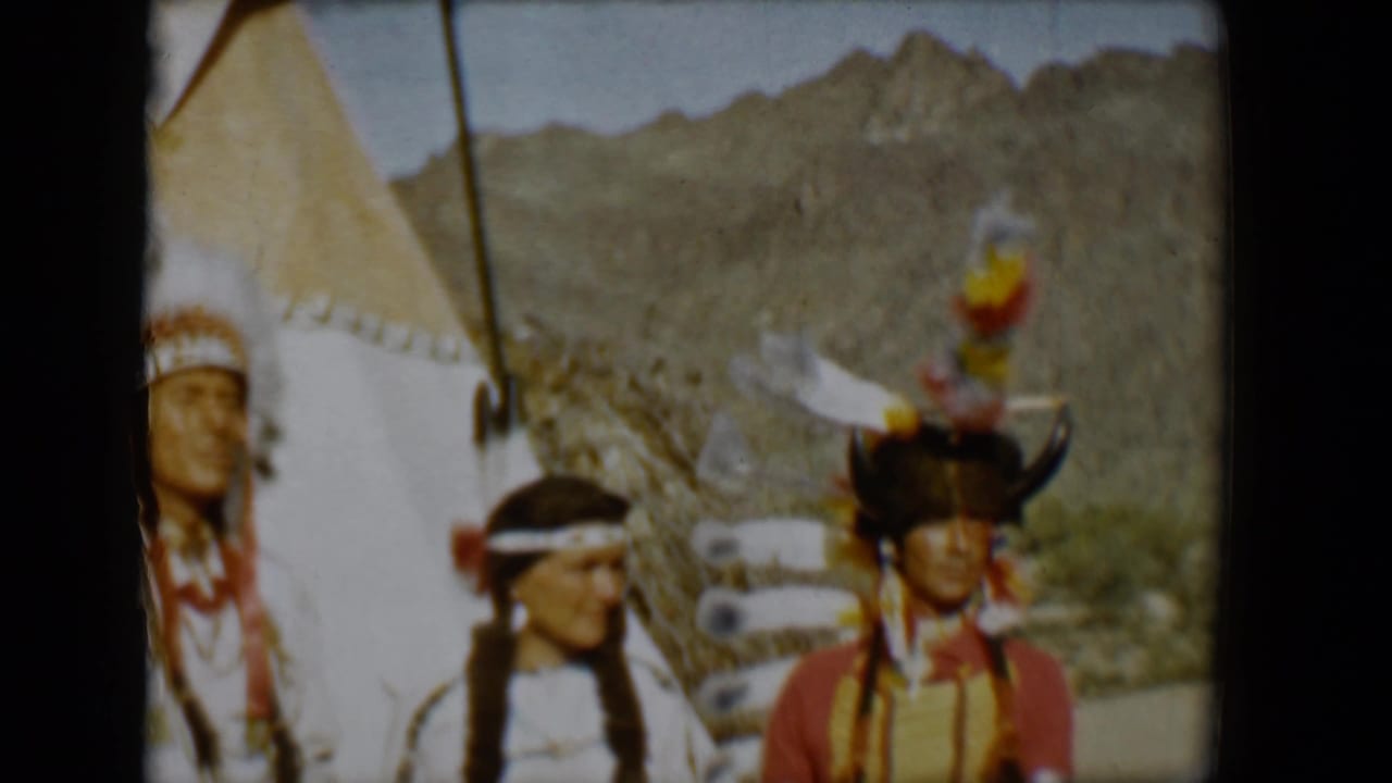 Three people in regalia pose near a teepee, with a mountain ridge in the background.