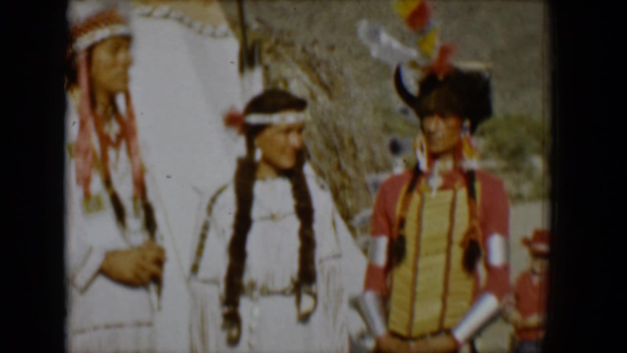 Three adults in Native American regalia stand near a teepee in a desert setting.