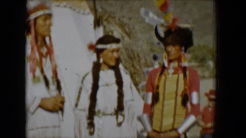 1958 Tucson home-movie frame showing adults in feathered headdresses and regalia near a teepee, with desert hills behind them.